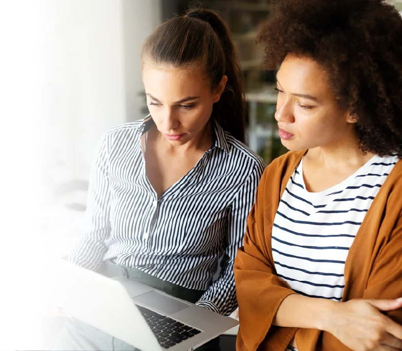 Two women working together and looking at a laptop screen in a bright office setting.
