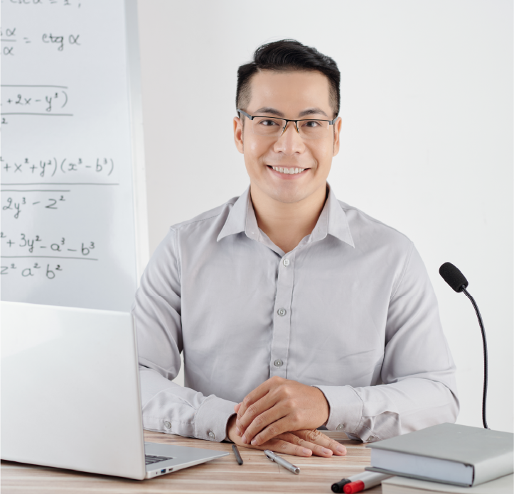 A smiling male professor sitting at a desk with a microphone, laptop, books, and a whiteboard behind him.