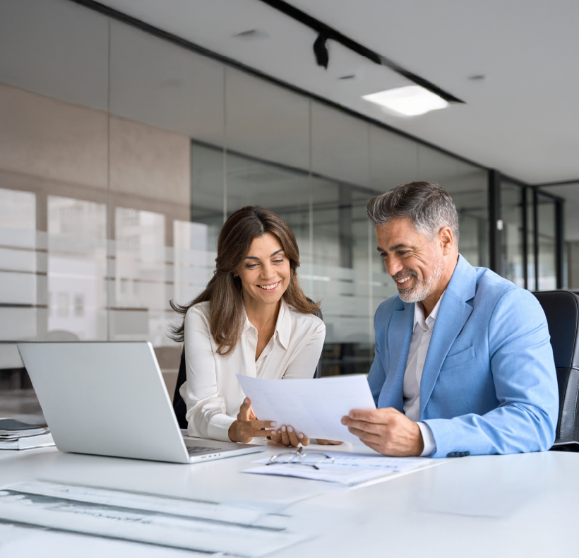 Two professionals reviewing financial documents and laptop data in a modern glass-walled conference room.