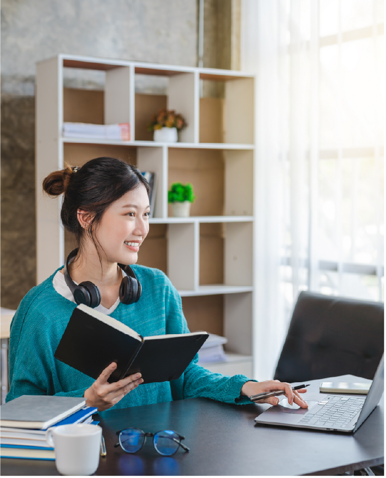 "A female student with headphones around her neck holds a notebook and smiles while using a laptop"