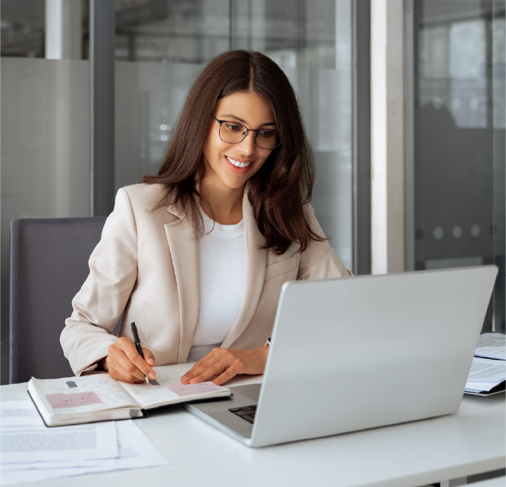 A professional woman smiles at her computer screen while preparing to take notes in a modern office setting.