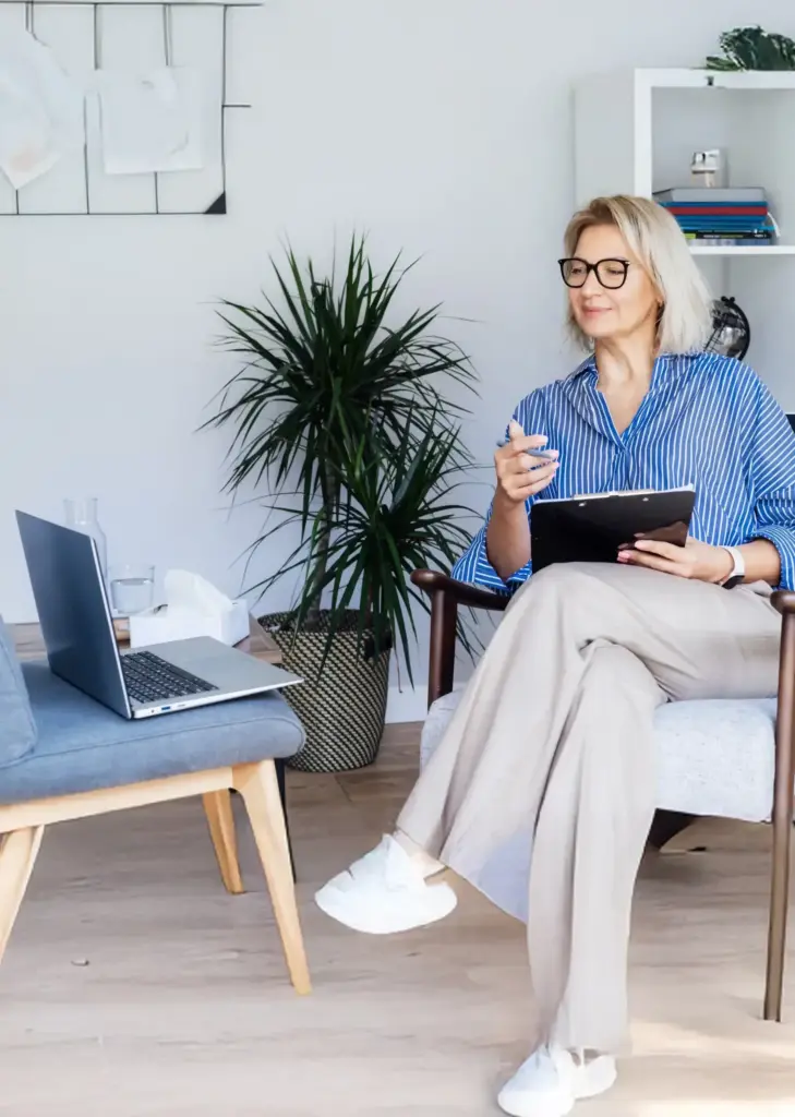 A woman sitting on a chair with a clipboard while looking at her laptop on another chair.