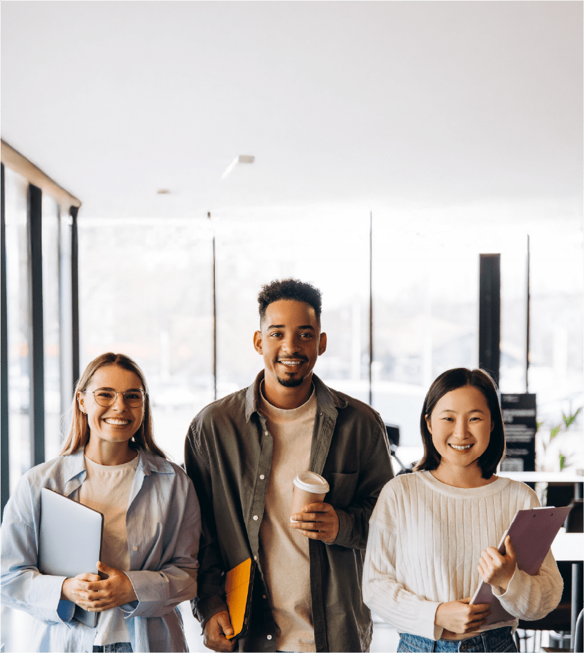 Three students smiling standing together, each holding a tab, a coffee and a file.