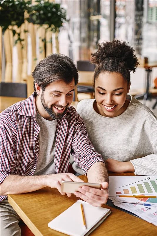 Two colleagues smiling while reviewing information on a smartphone beside printed charts and a notebook.
